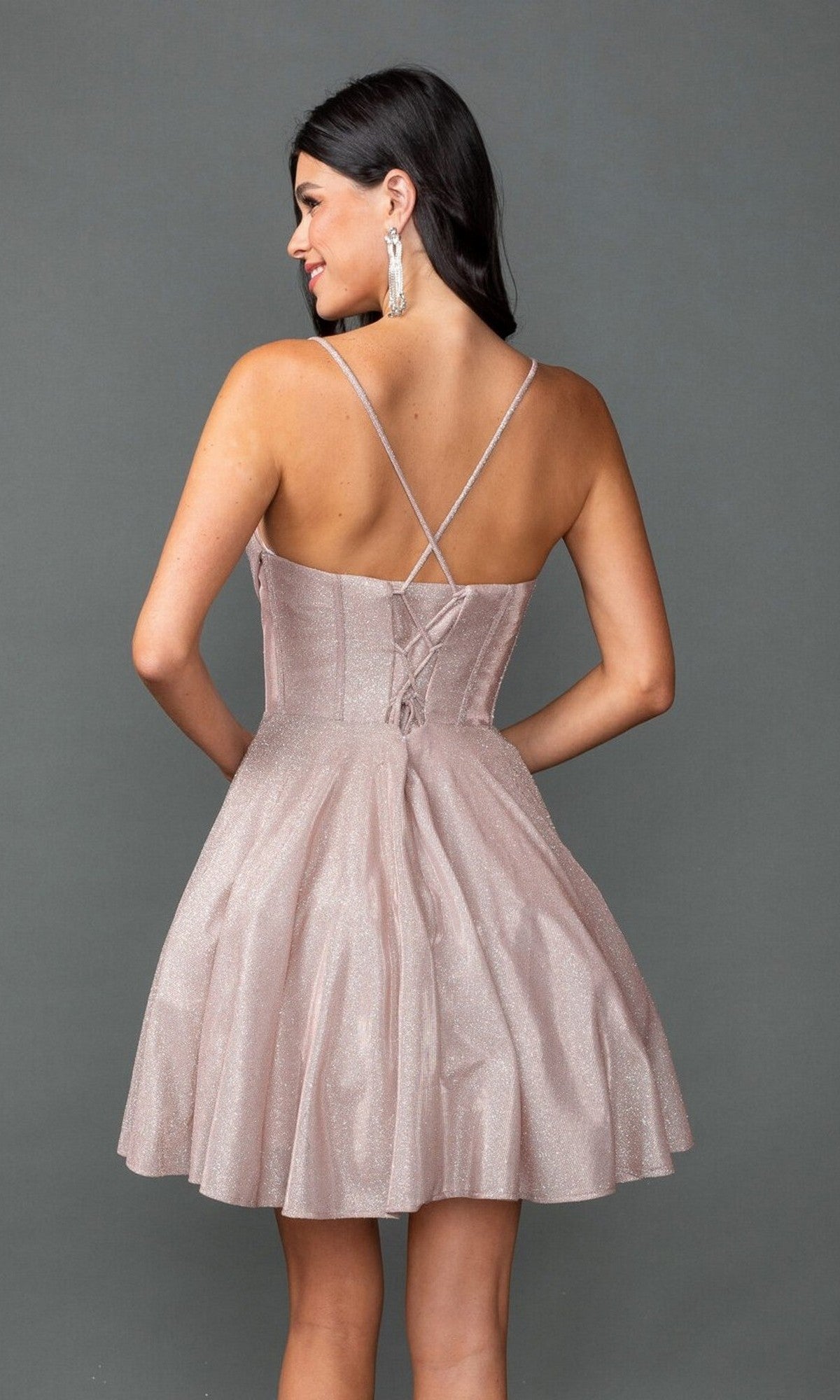 A woman wears the Dancing Queen 3421 short prom dress—sparkly light pink glitter, A-line skirt, sleeveless, with crisscross straps and a lace-up back—standing against a plain gray background.